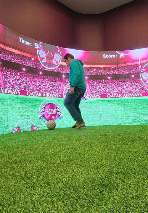 Person in a green hoodie plays soccer on a small artificial turf field indoors, with a large interactive curved screen displaying a stadium background, scoreboard showing Time: 90, Score: 2, and Qatar Airways logo.