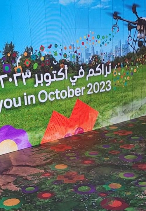 A digital interactive floor and wall display shows colorful flowers, a drone, and a city skyline with the text See you in October 2023.