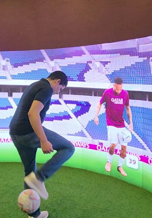 A man balances a football on his foot in front of a large interactive curved screen. The screen displays a football stadium with a player interacting with the man.