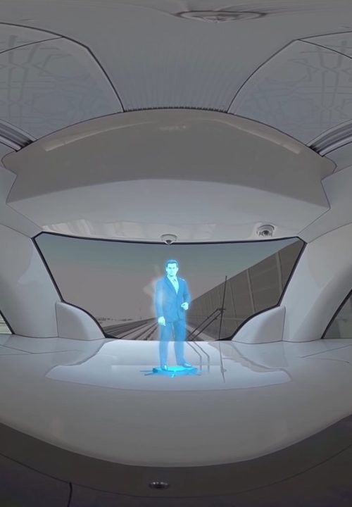 A futuristic train interior with sleek white design features a small blue hologram of a person standing near the front window, which shows a blurred cityscape view outside.