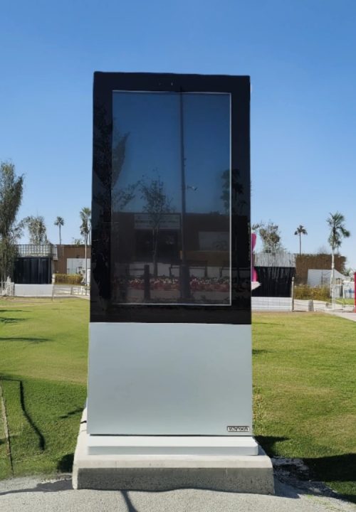 A large, vertical digital display stands on a concrete base in an outdoor park setting. The screen is turned off, reflecting the sky and surrounding trees. The area is sunny, with green grass and a clear blue sky in the background.