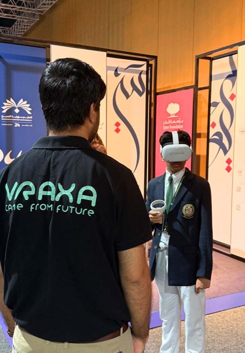 A person wearing a black VRAXA shirt observes another person using a VR headset at an exhibition. The display panels in the background feature intricate designs and the UNESCO logo.