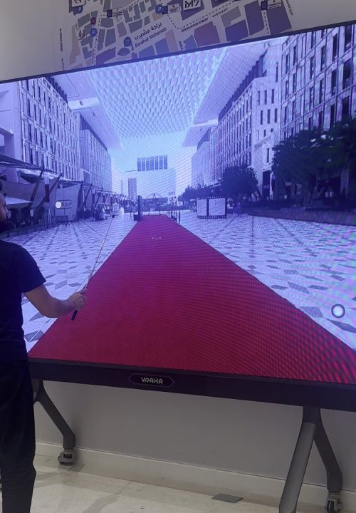 A person is interacting with a large interactive touch screen display that shows a virtual cityscape with a red carpet. They hold a long pointer stick, engaging with the content. The surrounding environment appears to be a modern indoor space.