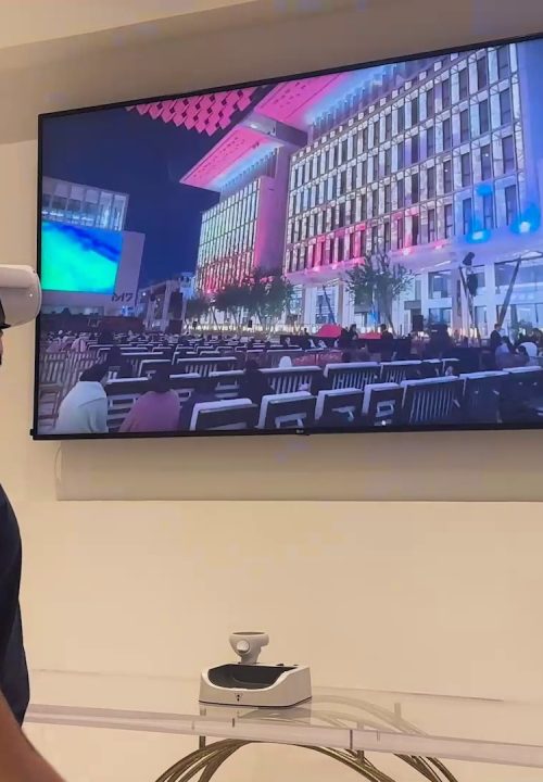 A person wearing virtual reality headset stands in front of a large wall-mounted TV casting an image of a building lit with colorful lights and a large audience seated outdoors. A gaming console is on a table below the TV.