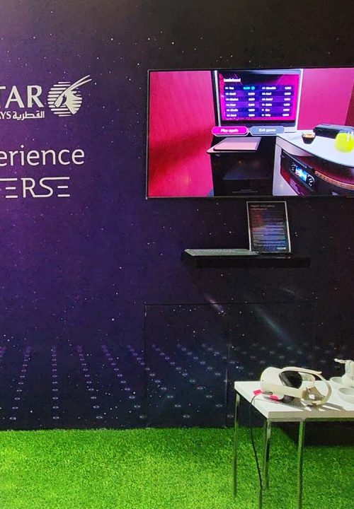 A Qatar Airways booth displays a screen and virtual reality headset on a small table on a grassy floor. The wall reads Experience Qverse with the airline logo. The screen shows a cabin interior, indicating a virtual experience.